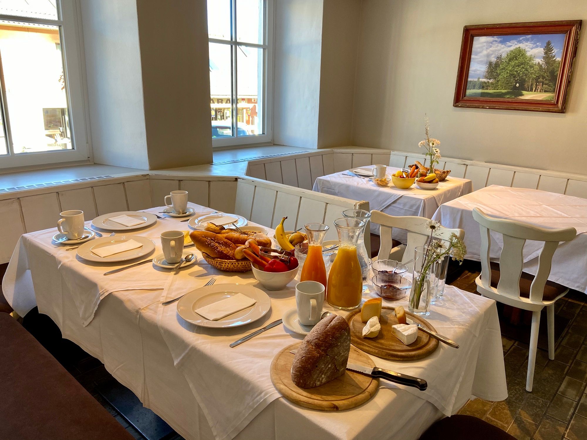 A laid breakfast table with bread, rolls, fruit, juice and crockery in a cozy room with windows and a painting on the wall.