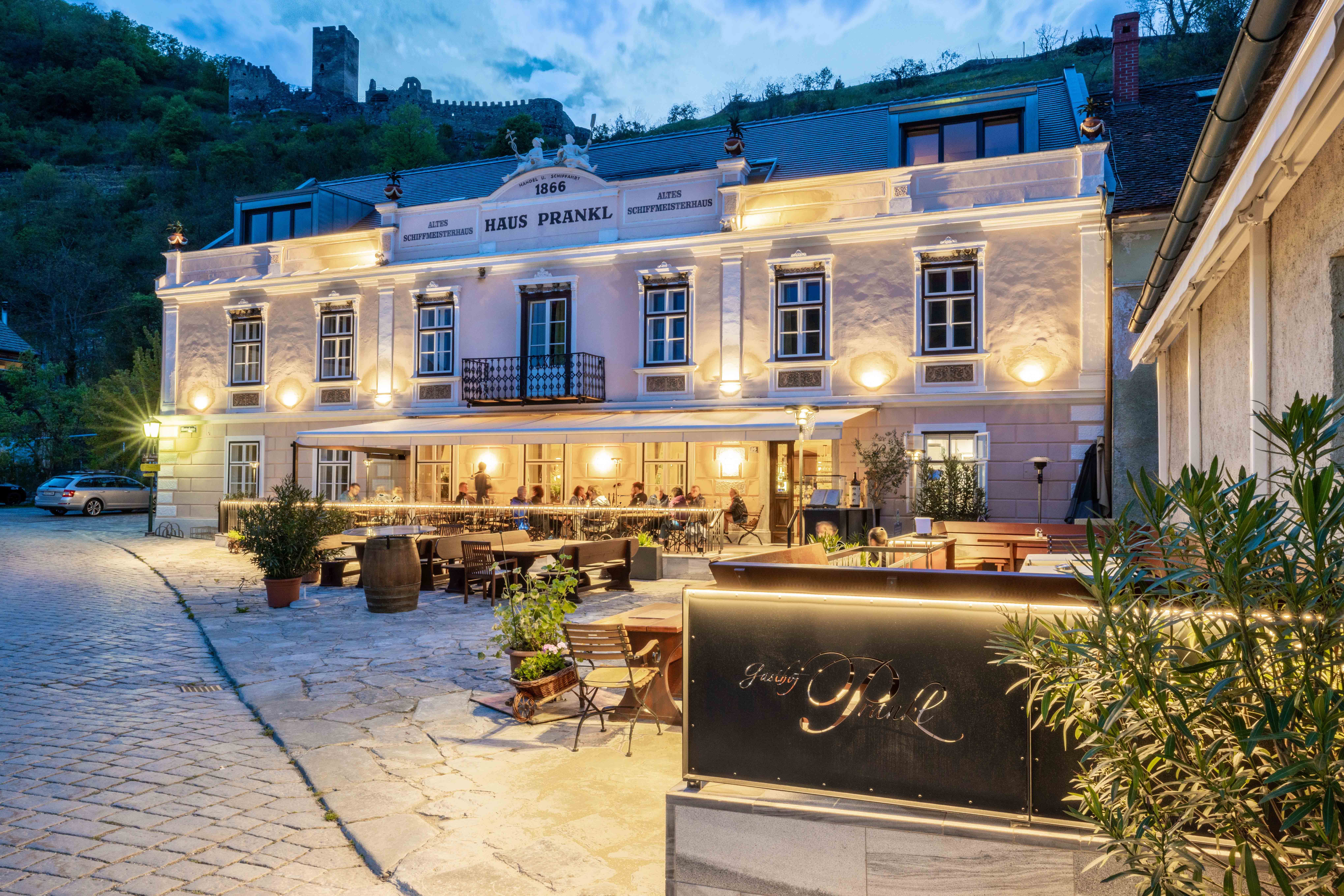 Exterior view of Gasthof Prankl at dusk with illuminated terrace and castle in the background.