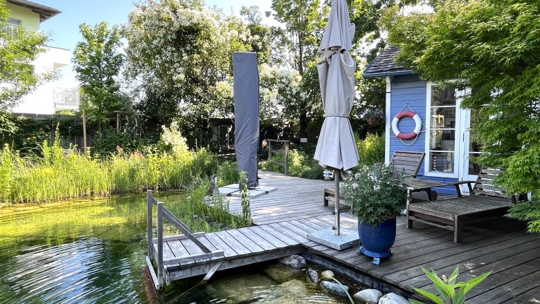 Garden with pond, wooden terrace, sun loungers and parasol.