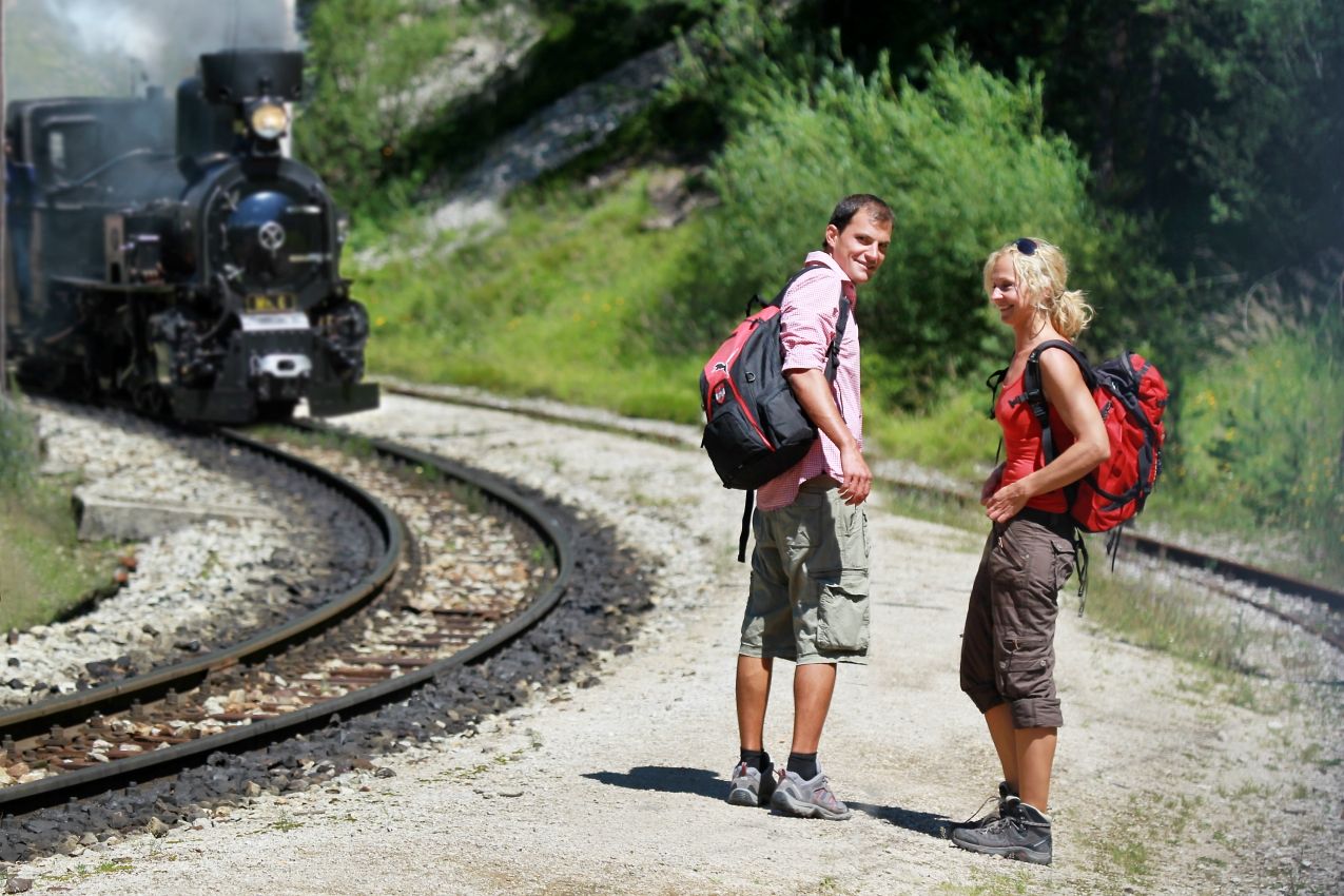 Two hikers stand on a gravel path next to railroad tracks while a steam locomotive passes by.