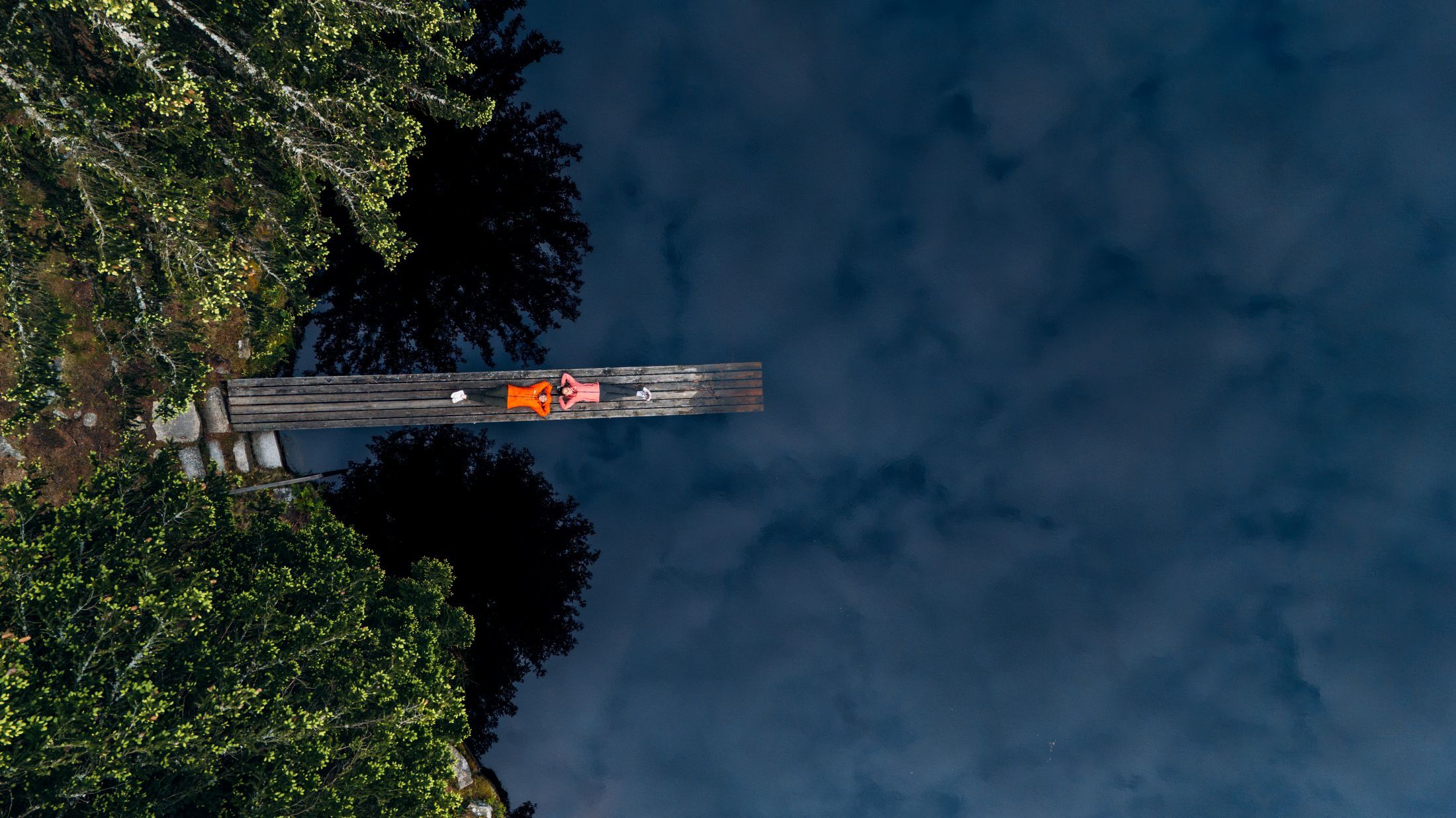 Two people are lying on a footbridge over a dark pond, surrounded by trees.