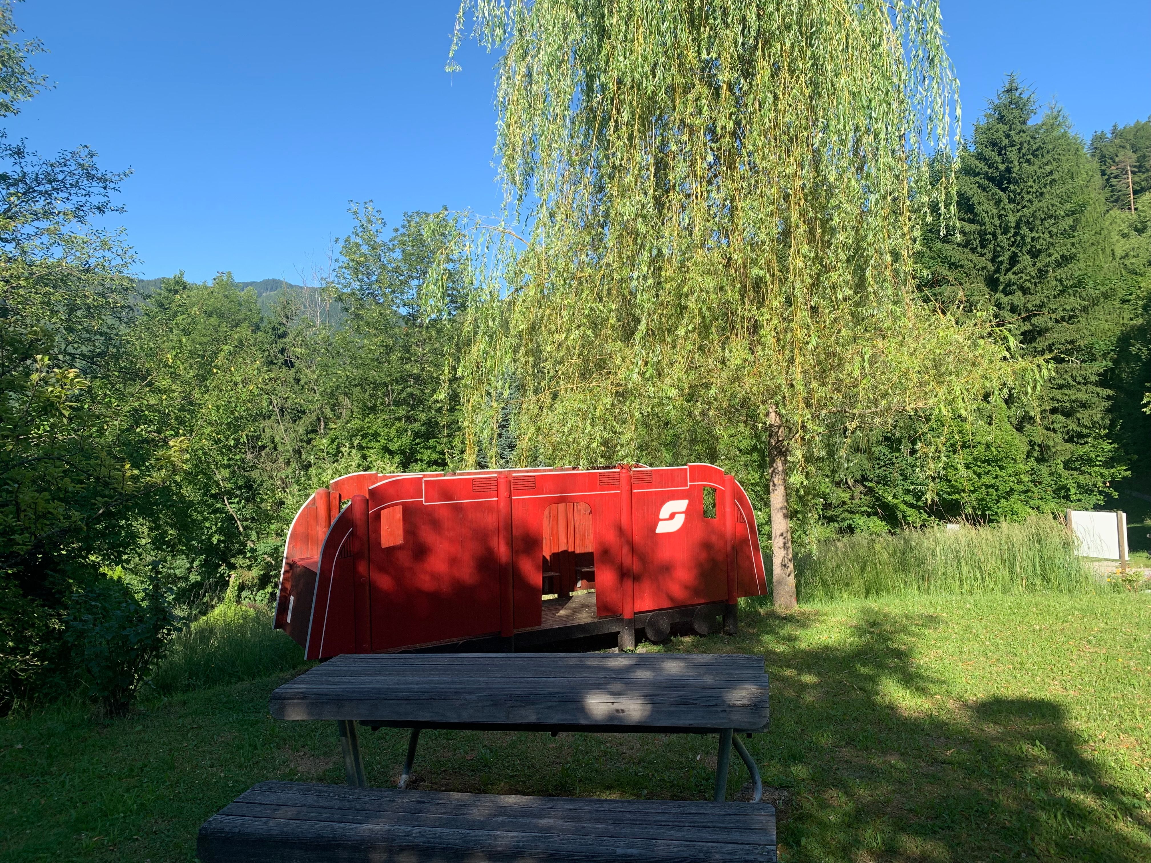 Red toy locomotive outdoors next to a tree and a wooden bench.