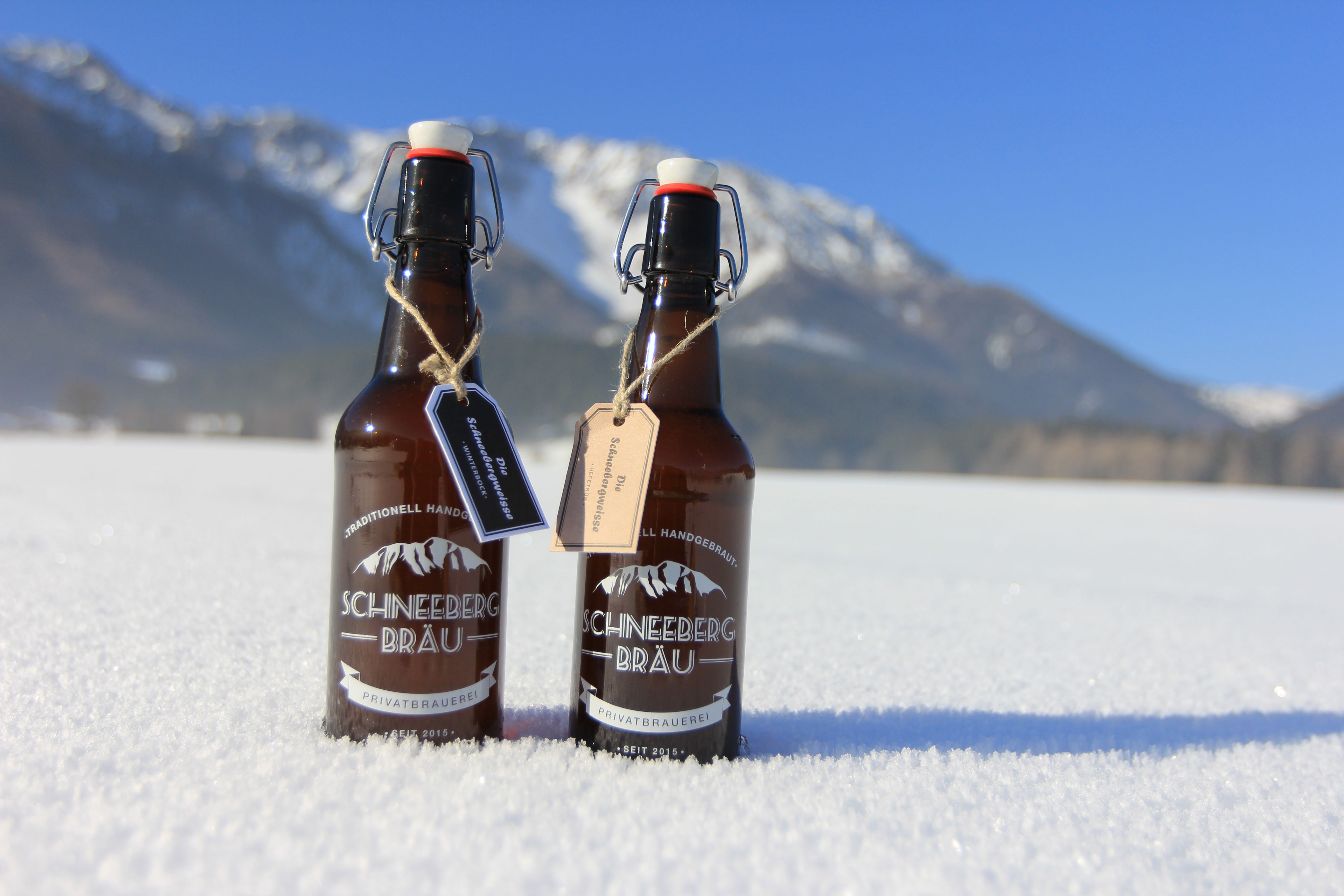 Two bottles of Schneeberg Bräu beer stand in the snow against a mountain backdrop.