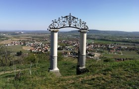 Two columns with an ornate arch on a hill overlooking a village and fields in the background.