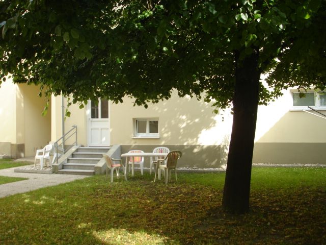 Inner courtyard with lawn, a tree and white plastic chairs. The yellow building in the background.