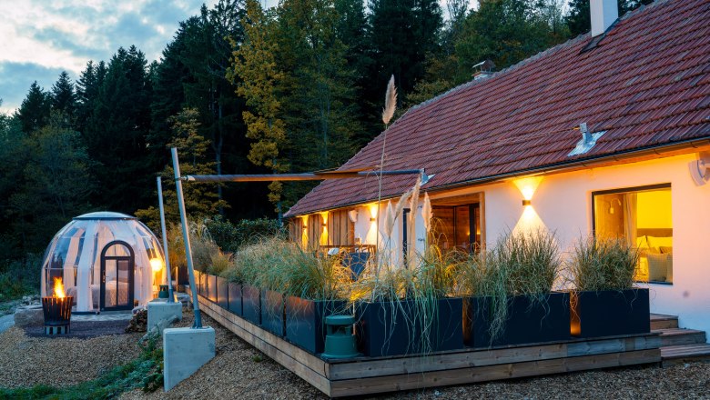 Chalets & places full of WONDER - Weidehaus De Luxe, © WUNDERVOLL eben A modern chalet with a terrace and a transparent dome tent in the forest at dusk.