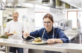 A chef in a restaurant kitchen prepares a dish while a colleague holds another dish in the background.