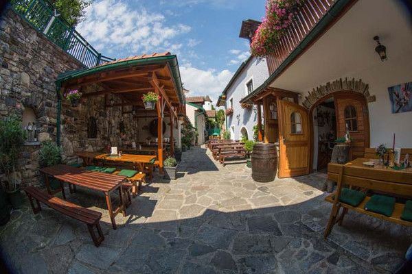 A cozy courtyard with wooden benches, tables and flowers. Stone floor and covered seating areas. Sunny weather with blue skies.
