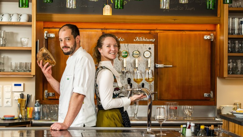 Two people are standing in a bar, one holding bread, the other a tray of drinks.