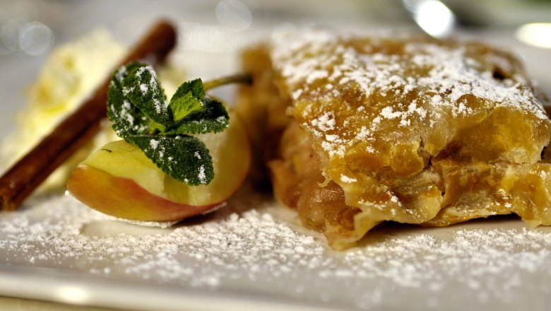 A piece of apple strudel with powdered sugar, apple slice, cinnamon stick and mint leaf on a plate.
