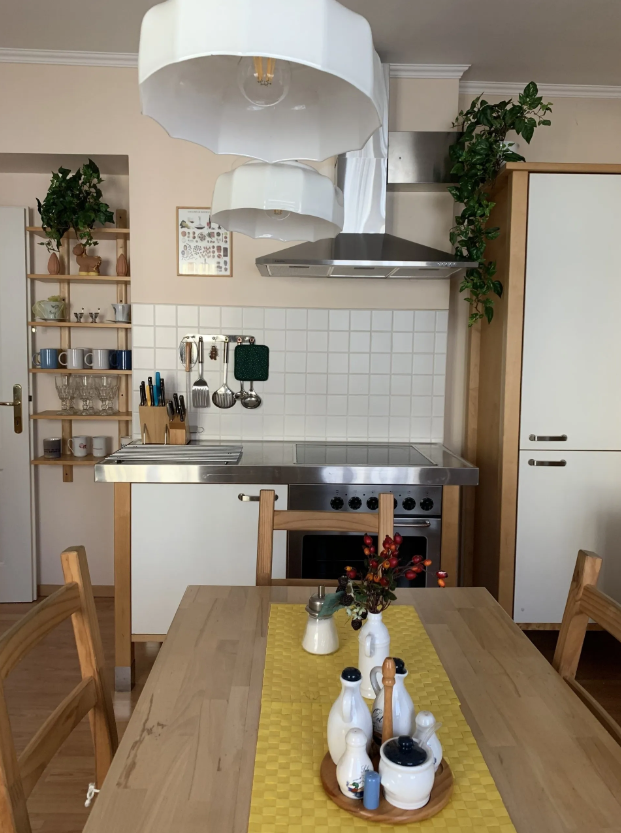 Cozy kitchen with wooden furniture, dining table and chairs, two windows with curtains, and kitchen utensils on the worktop.