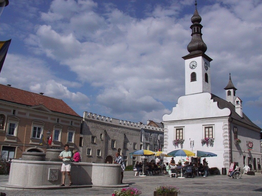 A town square with a white building with a tower and clock, people sitting under parasols, a fountain in the foreground.