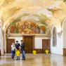 Interior view of a magnificent room in Göttweig Abbey with frescoes on the ceiling and three people talking.
