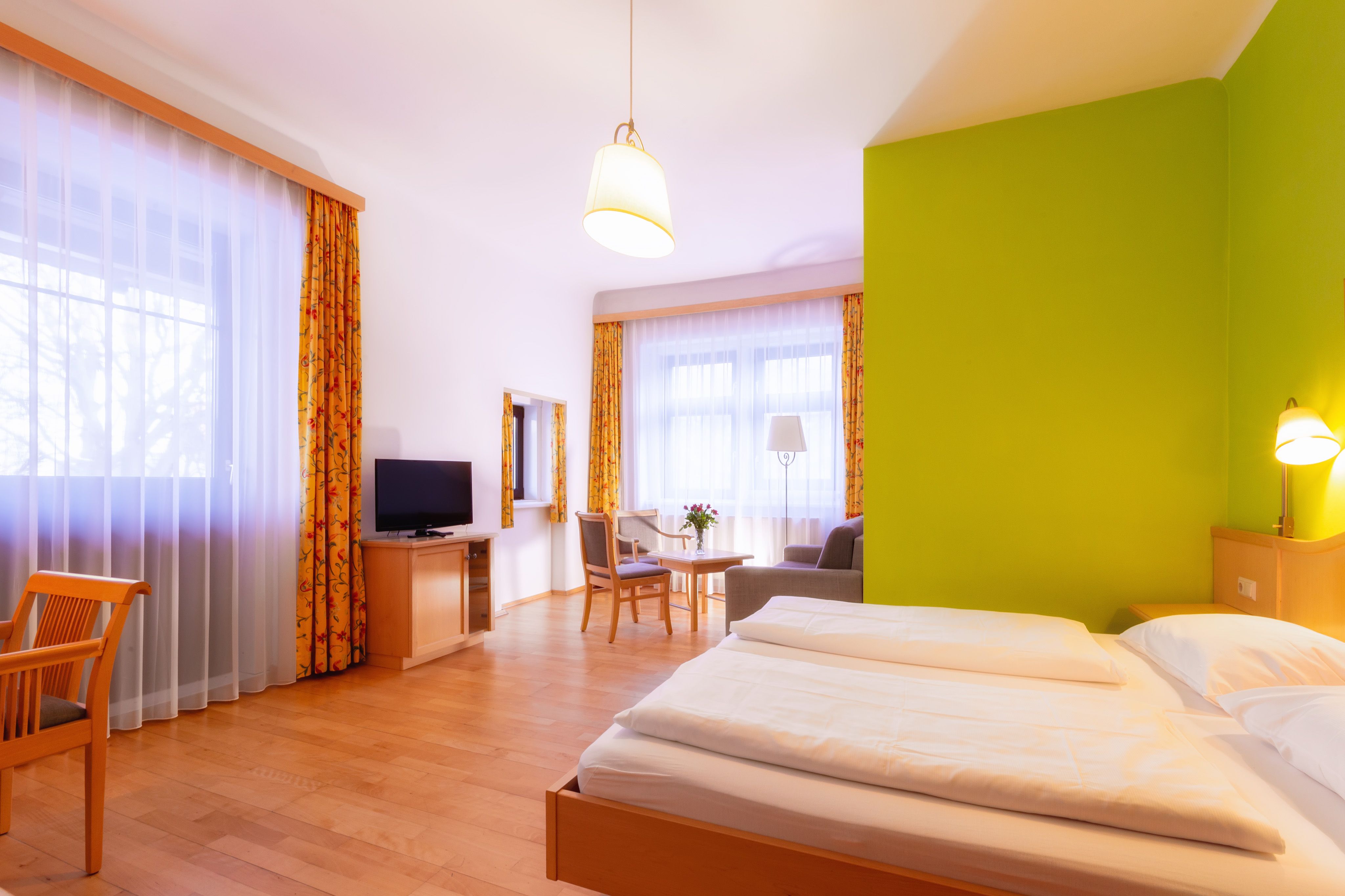 Bright hotel room with double bed, green accent on the wall, TV, seating area and large windows with curtains.