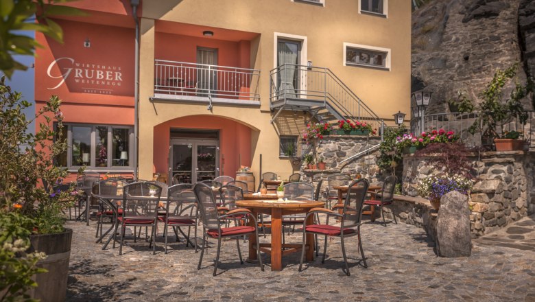 Outdoor area of a restaurant with tables and chairs on a paved courtyard, surrounded by flowers and plants.