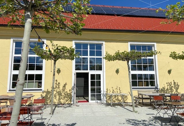 Yellow building with red roof tiles, large windows and a beer garden with tables and chairs outside.