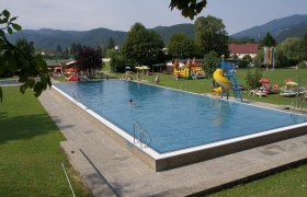 An outdoor pool with swimming pool, slide and sunbathing lawn, surrounded by trees and mountains.