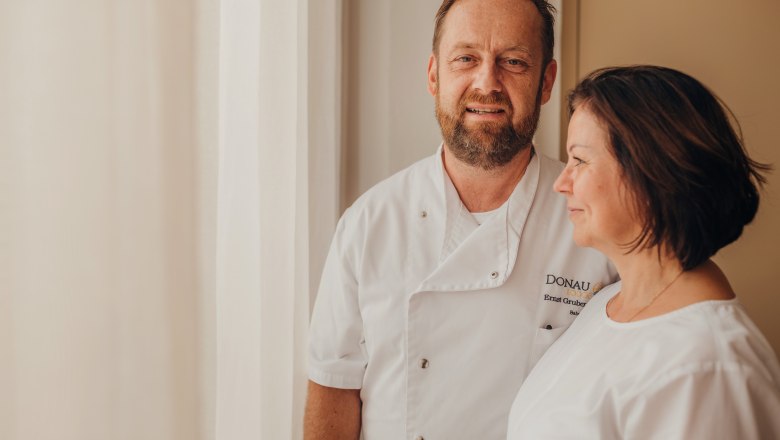 A man and a woman in white chef's jackets stand next to each other in front of a window.