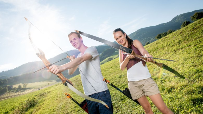 Two people doing archery in a meadow with mountains in the background.
