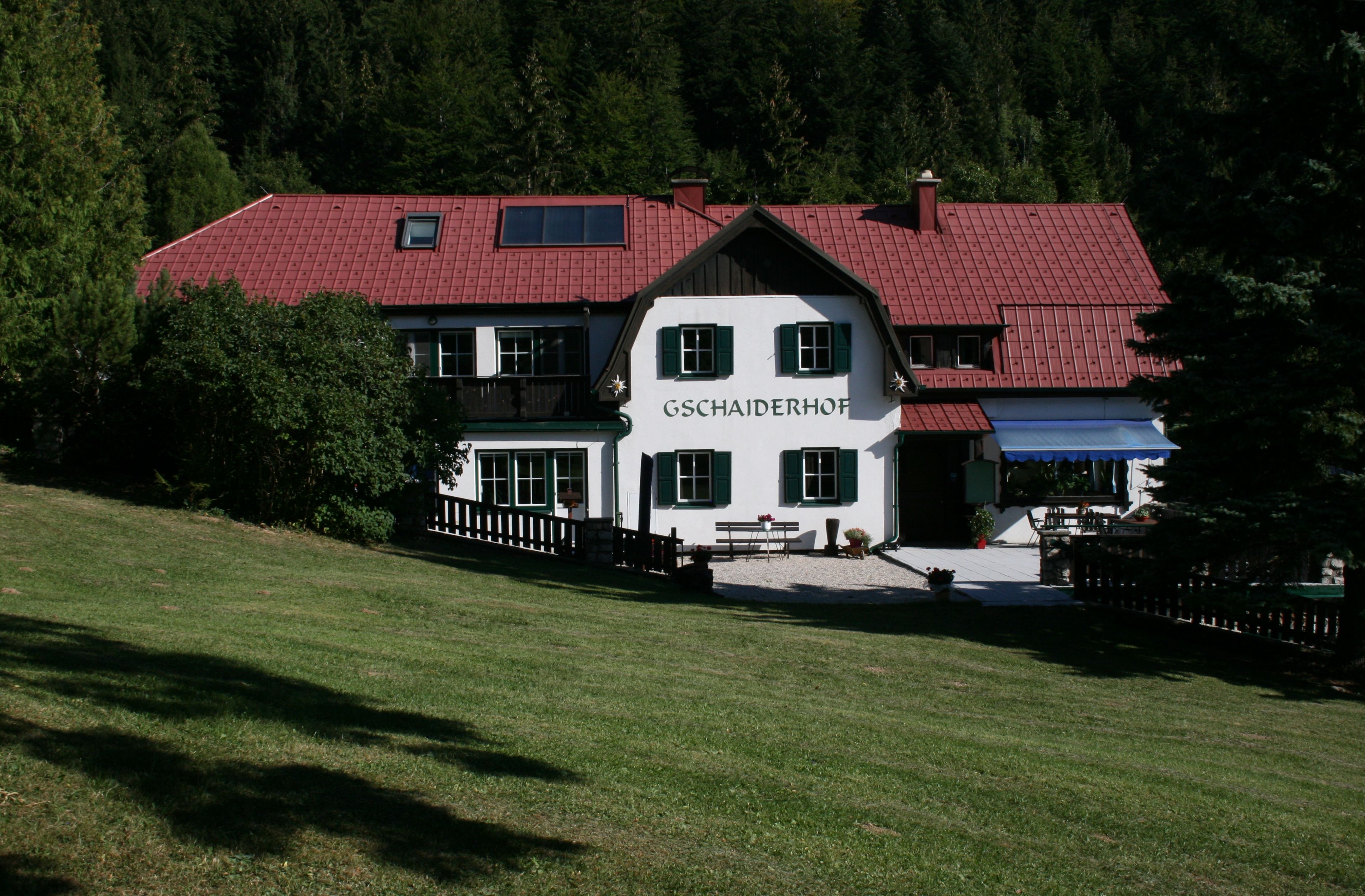 A traditional building with a red roof and green shutters, surrounded by trees and meadows, with the lettering 'Gschaiderhof'.