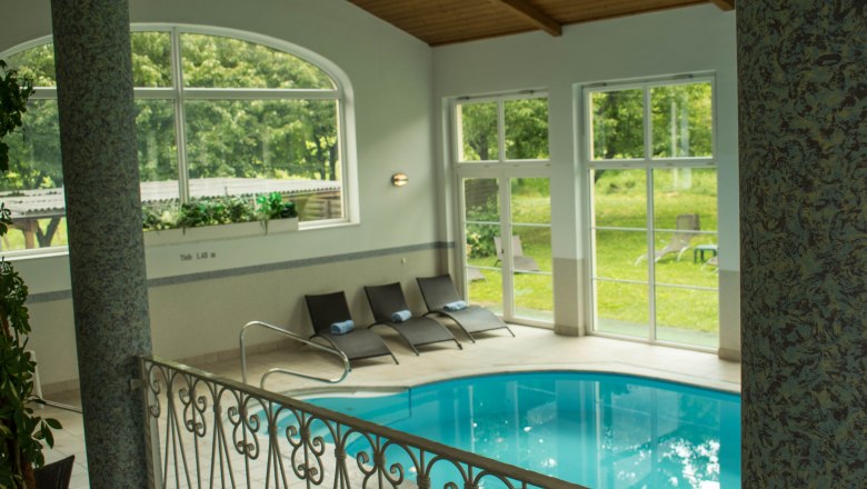 Interior view of a hotel pool with sun loungers and large windows overlooking the greenery.
