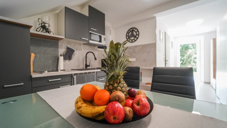 Modern kitchen with fruit bowl on the table, filled with pineapples, apples, oranges and bananas. Dark cupboards and light-colored walls in the background.