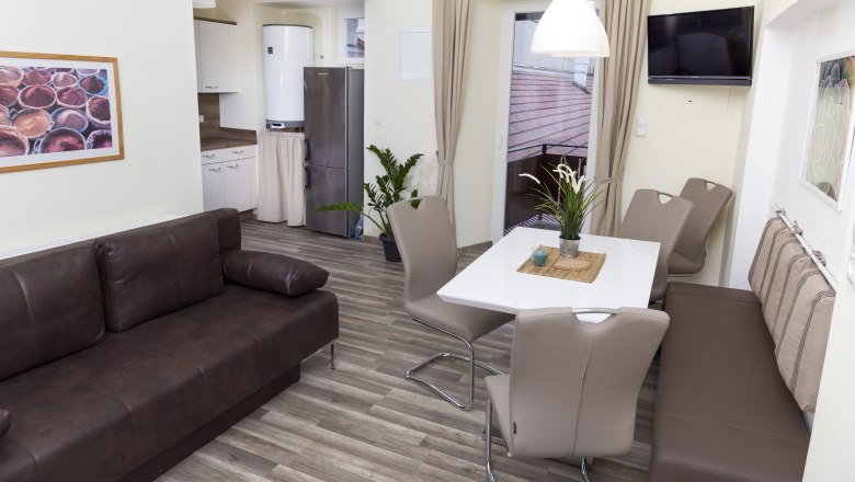 Modern kitchen with brown sofa, dining table and chairs, fridge and plants.