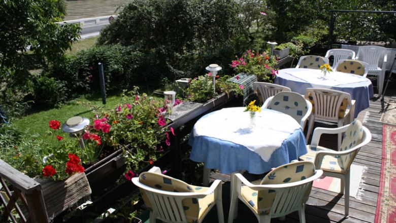 Terrace with tables and chairs, surrounded by plants and flowers, overlooking a river.