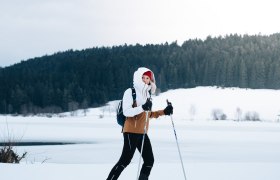 A person cross-country skiing on a snow-covered field in front of a forest.