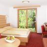 Bright hotel room with double bed, red carpet, table with fruit bowl and TV, view of green trees through a large window.