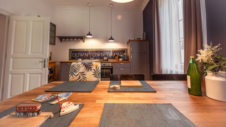 Kitchen with wooden table, toy train, bottle and flowers on the table, modern lamps and kitchen cabinets in the background.