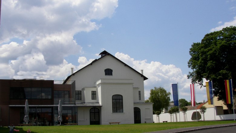 A modern concert hall with white walls and large windows, surrounded by flags and trees, under a cloudy sky.