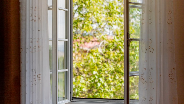 Open window with white curtains, view of green trees.