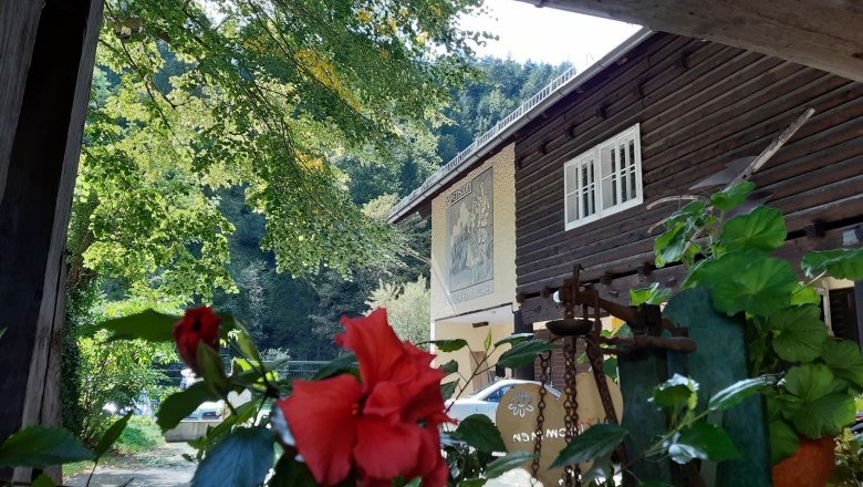 Idyllic courtyard, © Familie Kellner A traditional wooden house with red flowers in the foreground and green trees in the background.