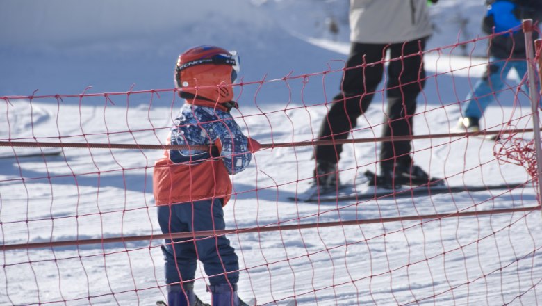 A child with a red helmet and ski equipment stands behind a red safety net on a ski slope.