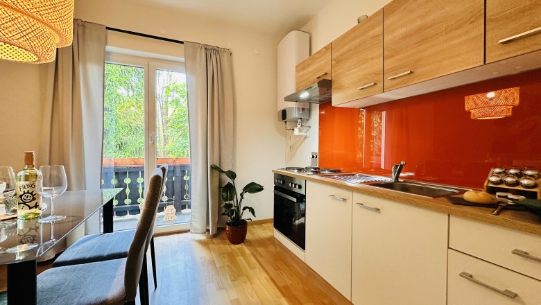Cozy kitchen with wooden furniture, red splashback and dining table. View of balcony with plants.