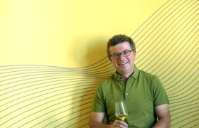 Man with glasses and green shirt holds a wine glass in front of a yellow background with a line pattern.