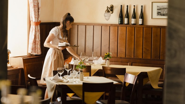 A woman in traditional dress sets a table in a cozy restaurant with wooden walls and bottles of wine in the background.