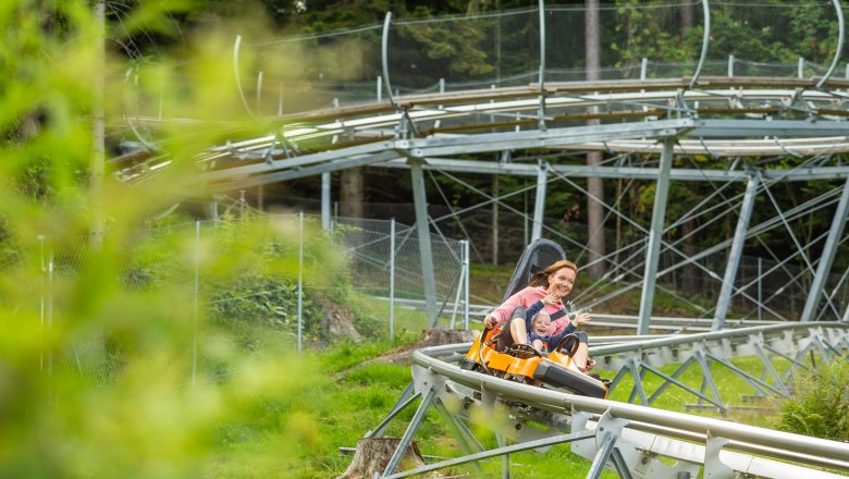 A woman and a child ride through a green landscape on a summer toboggan run, laughing.
