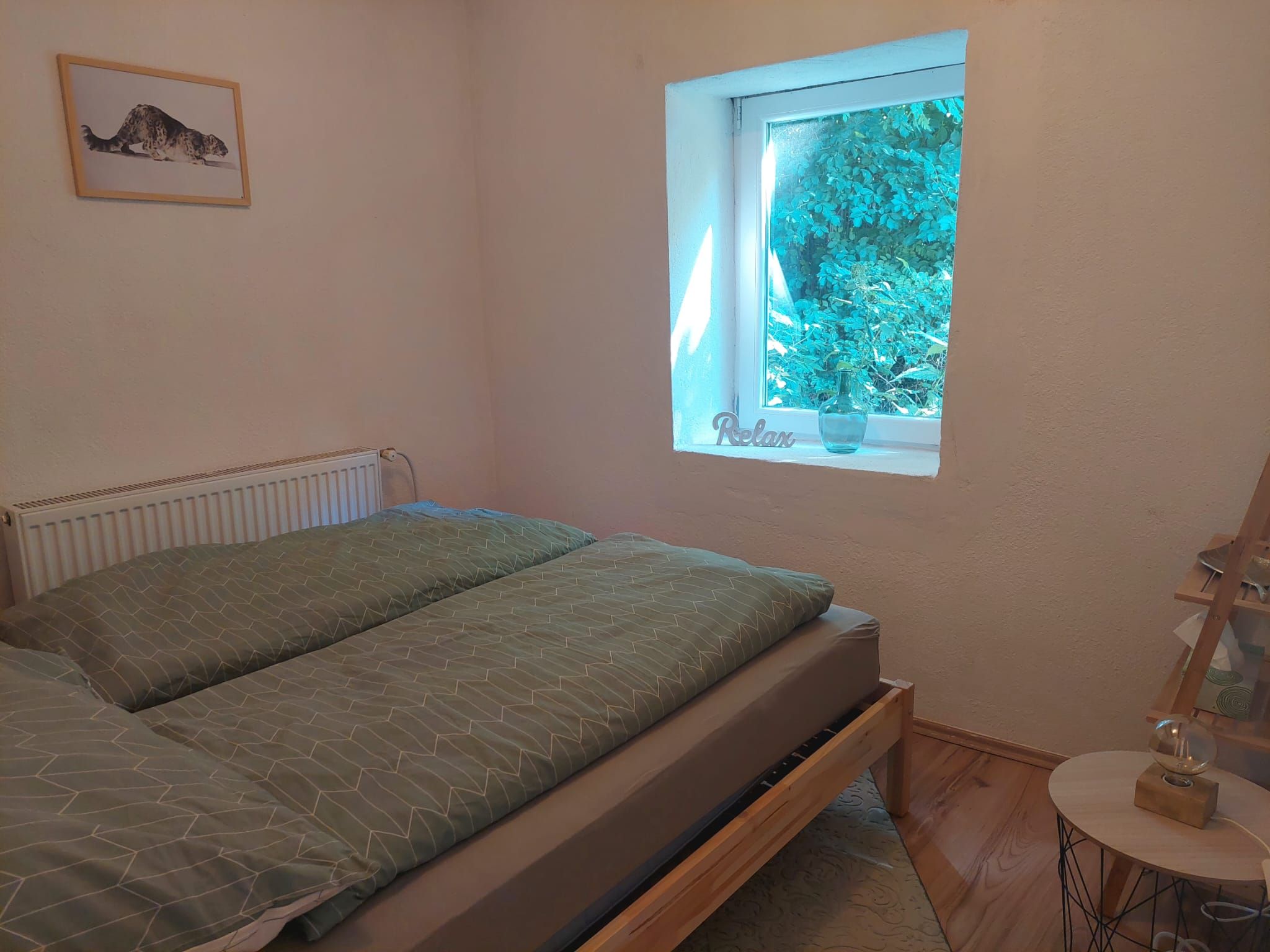 A cozy bedroom with a bed, a window with a view of the greenery and a picture on the wall.