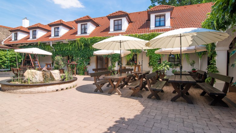 A cozy courtyard of a winery with wooden tables, benches and parasols, surrounded by a building with red tiled roofs and green vegetation.