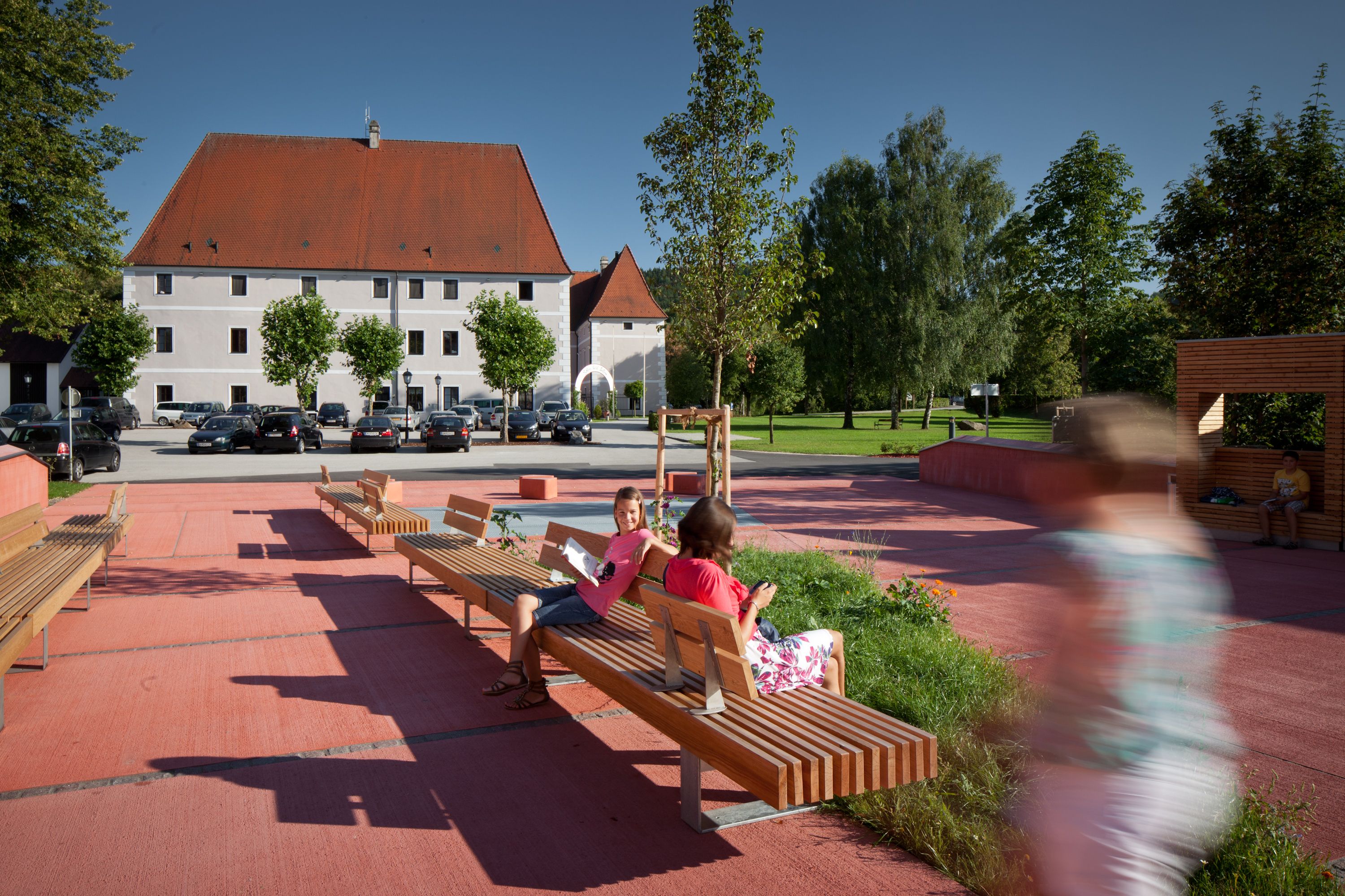 A modern square with wooden benches and people in a sunny setting, with a large building with a red roof in the background.