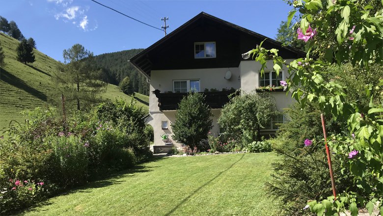 A two-storey house with a balcony, surrounded by a well-tended garden and green hills in the background.