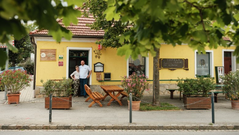 A yellow inn with a red roof, surrounded by trees. Two people are standing in front of the entrance.