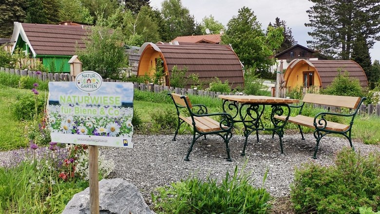 A sign with the inscription 'Naturwiese für Biene & Co' (Nature Meadow for Bees & Co) stands in front of a meadow with benches and tables, surrounded by small, wooden huts.