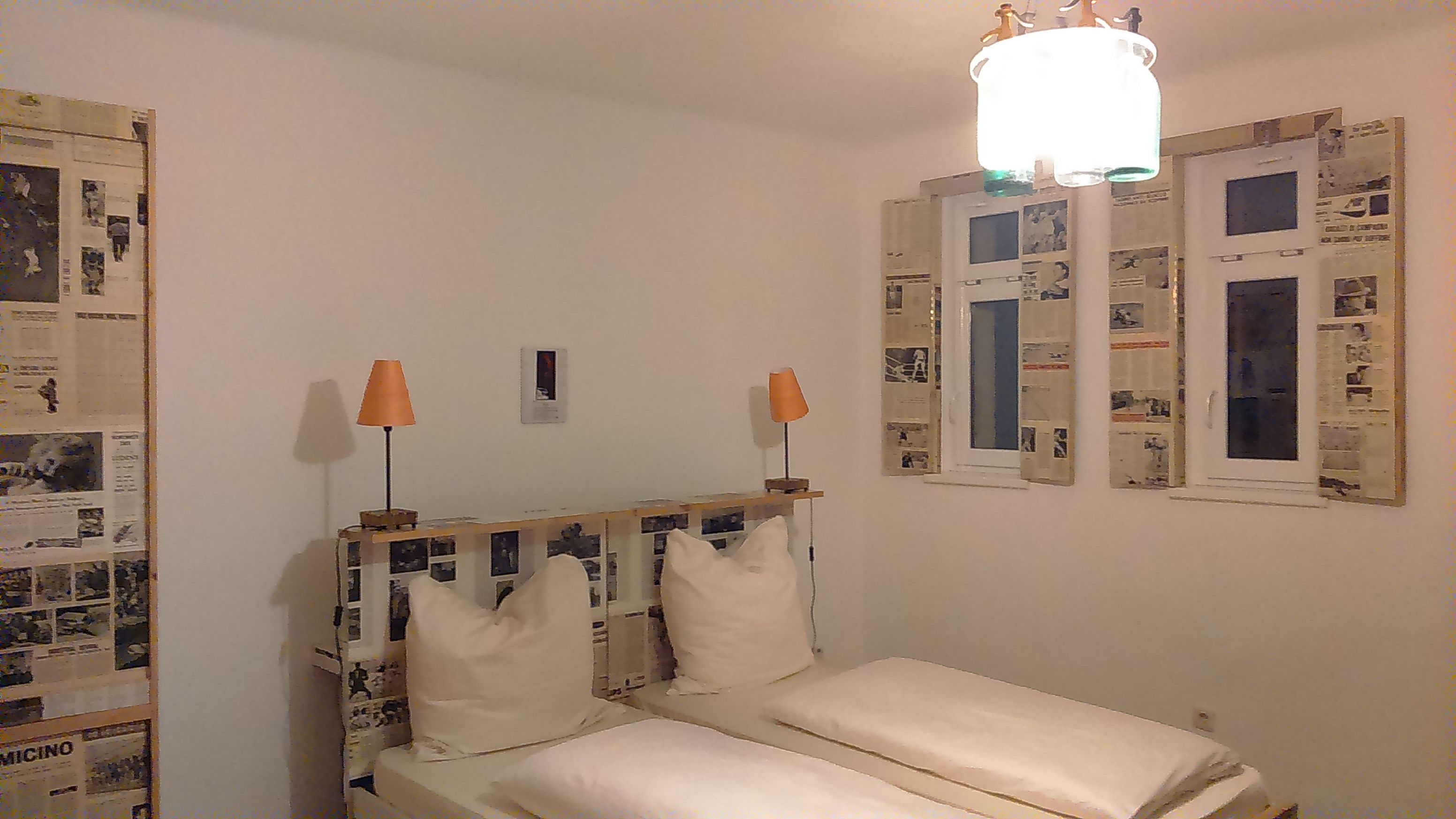 Hotel room with double bed, newspaper decorations on the walls and windows, two orange lamps.