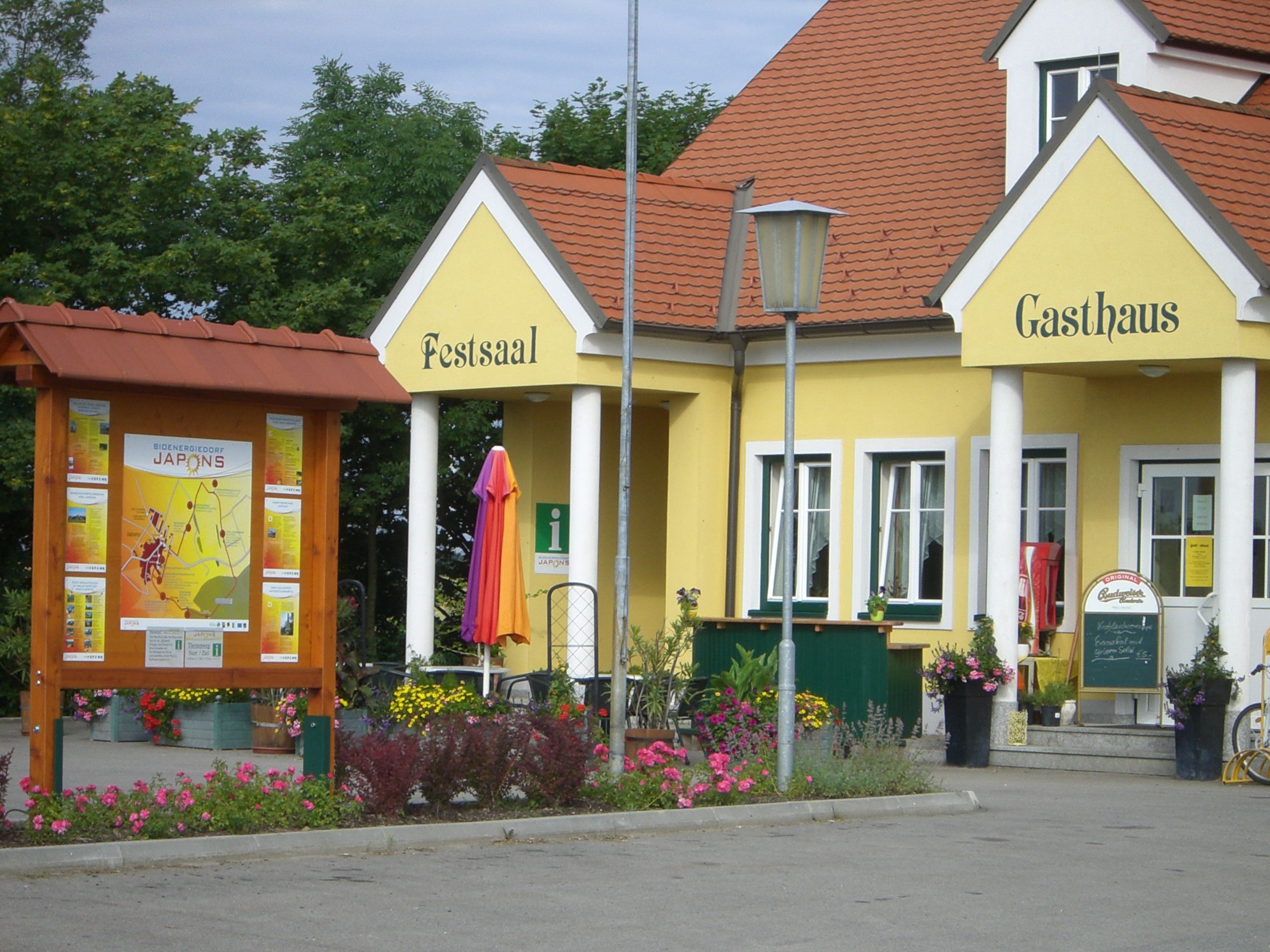 Yellow building with the inscription 'Festsaal' and 'Gasthaus', surrounded by flowers and an information sign.