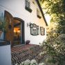 Entrance to a country house with white walls and green shutters, surrounded by flowering plants.
