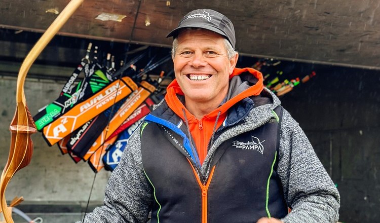 A smiling man holds a bow in front of a pendant with arrows.