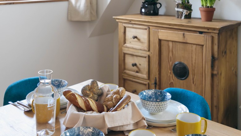 A laid breakfast table with bread rolls, crockery and yellow cups, a wooden cupboard with plants in the background.